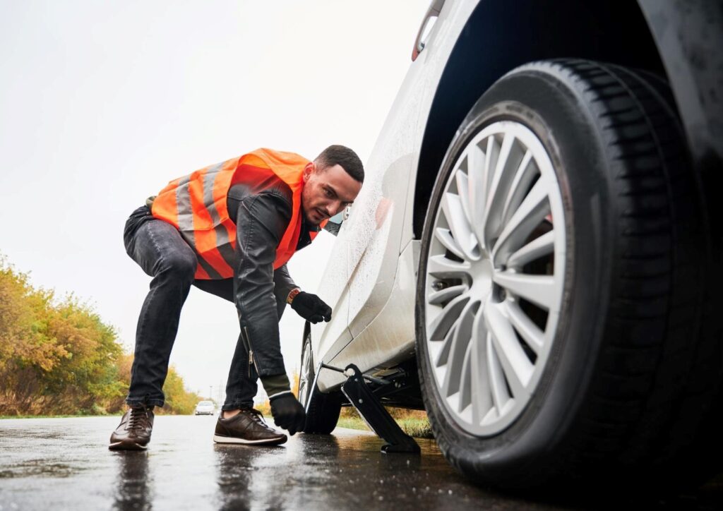 Man in a bright orange safety vest changing a car tire on a wet road, using a jack.