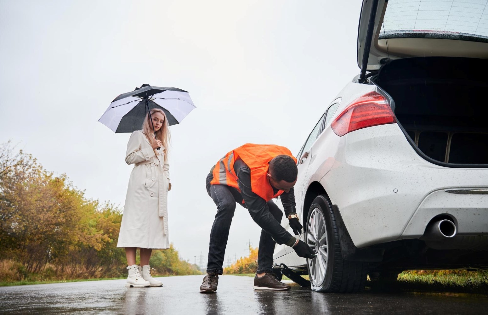 Roadside Assistance Lexington KY | Metro Towing | Metro Towing Woman in a beige trench coat stands with a black-and-white umbrella as a man in an orange safety vest changes a flat tire on a white car on a wet autumn road behind her.
